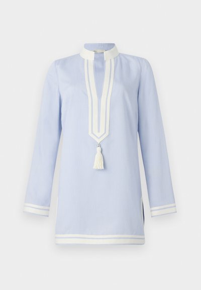 Gaiši zila tunika ar garām piedurknēm, kurai ir balts apdarināts apkakle, dekoratīvas baltas svītras un pušķa detaļa ap kaklu.
