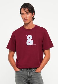 Vīrietis ar tumšiem matiem valkā maroon krāsas T-kreklu ar baltu simbolu "&" un tekstu "me" augšpusē un "my calvins" apakšpusē, apvienojot to ar izbalējušām pelēkām džinsām.