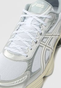 White mesh running shoe with silver synthetic overlays, featuring a textured pattern, flat white laces, and a beige outsole.