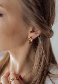 Goldene Creolen, die in einem gepiercten Ohr getragen werden, mit einer glatten, polierten Oberfläche. Die Haare sind locker mit einer leichten Welle frisiert.