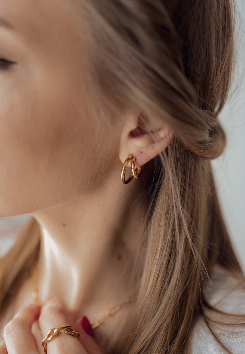 Goldene Creolen, die in einem gepiercten Ohr getragen werden, mit einer glatten, polierten Oberfläche. Die Haare sind locker mit einer leichten Welle frisiert.