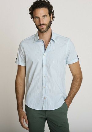 Homme aux cheveux bouclés et à la barbe portant une chemise bleu clair à manches courtes boutonnée et un pantalon vert, debout avec une main dans la poche.