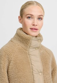 Sandfarvet jakke uden krave med en blød, pelsagtig tekstur på ydersiden og en glat beige foring. Har to trykknapper.
