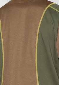 Suéter com a parte da frente em castanho e painéis laterais em verde, contornado com costura amarela. O material parece texturizado com um design casual.