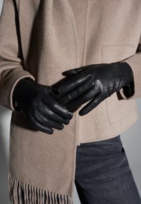 Schwarze Lederhandschuhe mit sechs Fingerähnhten, glatter Textur, getragen von Händen, die auf einem beigen Wollmischmantel mit Fransen-Details ruhen.