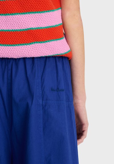 Person trägt ein rot-rosa gestreiftes Strickoberteil und blaue Hose mit dezentem gesticktem Text auf der Tasche.