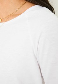 Weißes Baumwoll-T-Shirt mit einem leicht lockeren Schnitt, rundem Halsausschnitt und glatter Textur. Zarte goldene Halskette sichtbar.