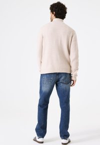 Helles beige, geripptes Strickpullover mit hohem Kragen, kombiniert mit ausgewaschenen blauen Jeans. Die Jeans verfügen über Gesäßtaschen und haben eine lockere Passform.