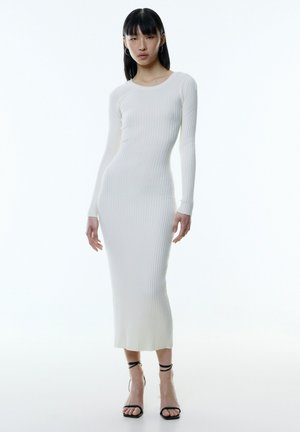 Langarm, aansluitende geribde jurk in off-white, met een ronde halslijn en midden kuitlengte, gecombineerd met zwarte bandjessandalen.
