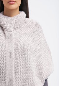 Poncho gris clair en maille avec un col haut, présentant un motif texturé et une fermeture par bouton au niveau du cou. Matière douce et confortable.