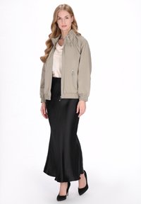 Beżowa kurtka bomberka z wysokim kołnierzem, elastycznymi mankietami i kieszeniami na zamek, zestawiona z czarną satynową spódnicą i butami na wysokim obcasie.