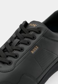 Sapatilha de couro preto com detalhes texturizados, logotipo dourado "BOSS" e atacadores pretos. Apresenta uma superfície lisa e um padrão perfurado na lateral.