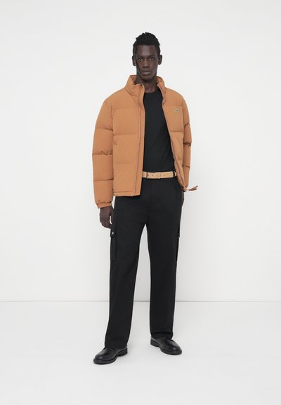 Veste matelassée marron avec un col haut, t-shirt noir, pantalon cargo noir et chaussures noires. Détails notables incluent un logo sur la veste.