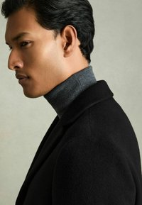 Sort frakke med høj krave over en grå turtleneck, der har en glat tekstur og skræddersyet design. Baggrunden er neutral og subtil.