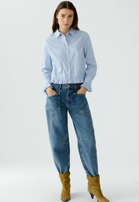 Hellblau gestreiftes Button-up-Shirt mit gerafftem Saum und langen Ärmeln; kombiniert mit lockeren, blauen Denim-Hosen und beigen Stiefeletten.