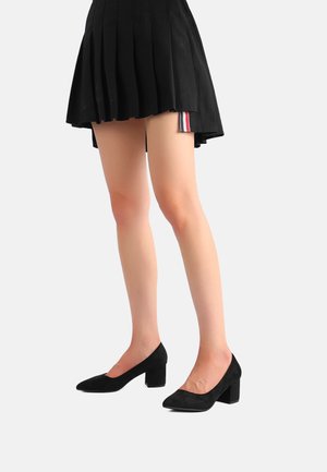 Scarpe con tacco alto in suede nero, con punta affusolata e tacco a blocco, indossate con una gonna nera plissettata. Texture liscia e design elegante.