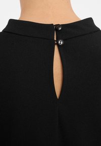 Schwarzes Stoffkleidungsstück mit Kragen, das hinten einen Schlüsselaugen-Ausschnitt und zwei schwarze Knopfverschlüsse hat. Weiche Textur und taillierte Passform.