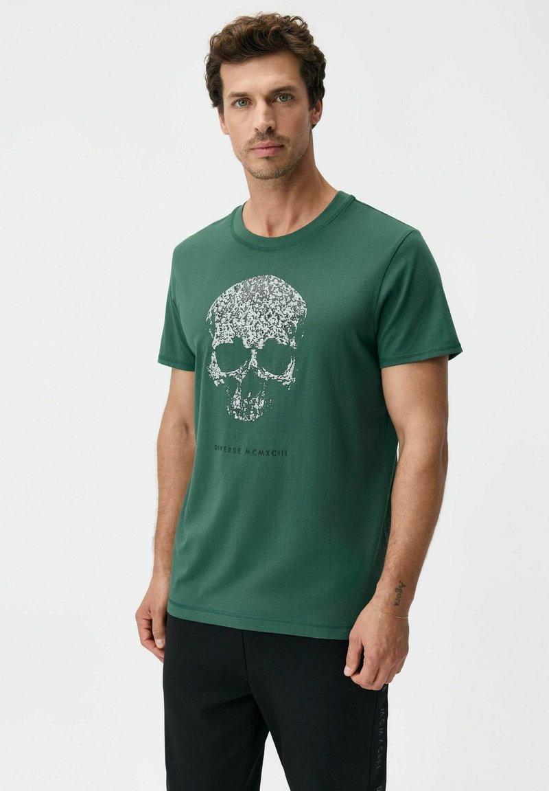 Zielony bawełniany T-shirt z krótkim rękawem z białym nadrukiem czaszki oraz napisem „Diverse MCMXCIII” poniżej grafiki. Luźny krój casual.