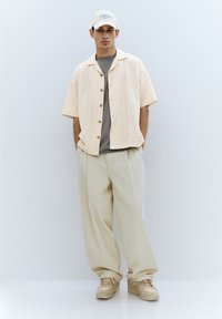 Short-sleeve beige button-up shirt with vertical texture, gray t-shirt underneath, cream trousers, and light sneakers. Cap with logo.