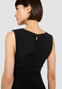 Juoda aptempianti suknelė su apvaliu iškirpimu nugaroje, raukšlėtu piešiniu palei stuburą ir subtilia aukso spalvos užtrauktuku užsegimui.