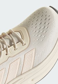 Svetlo béžová športová obuv s textúrovaným sieťovým vrchným materiálom, gumovou podrážkou a nenápadnými trojuholníkovými akcentmi. Šnúrky sú tenké a béžové.