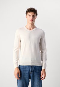 Maglione beige a V in maglia leggera, maniche lunghe e polsini a costine, abbinato a jeans in denim blu. Dettaglio del logo discreto sul petto.