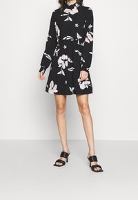 Musta kukallinen mekko pitkillä hihoilla, vyötäisiltä tiukennettu ja liehuva helma. Koristeena suuria valkoisia ja vaaleanpunaisia kukkia, yhdistettynä mustiin korkokenkiin.