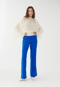 Maglione a coste crema con maniche a volantino, abbinato a pantaloni flare blu vibrante. L'outfit presenta eleganti scarpe bianche a punta.