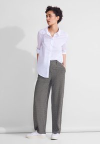 Lyhyttukkainen nainen, jolla on valkoinen napitettava paita ja kuviolliset leveälahkeiset housut, seisomassa käsivartensa taskuissa valkoista seinää vasten.