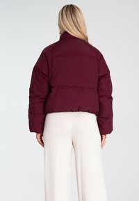 Burgundowa kurtka puchowa, z lekko skróconym krojem, pikowaną fakturą i wysokim kołnierzem, noszona na kremowych szerokich spodniach.