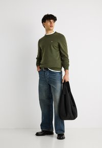 Jeune homme portant une chemise verte à manches longues et un jean bleu, tenant un sac noir dans sa main gauche, debout contre un mur blanc.
