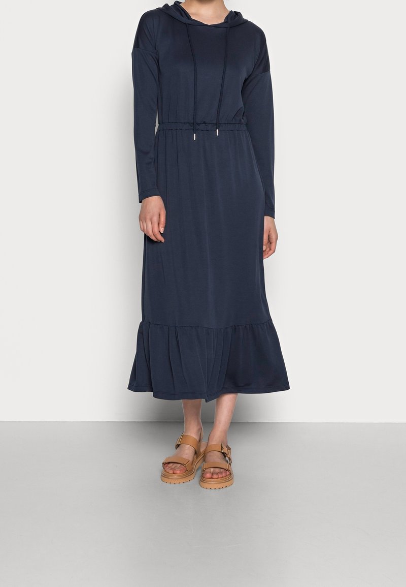 Femme portant une robe midi manches longues bleu marine avec une capuche et une taille à cordon, associée à des sandales compensées couleur tan.