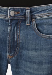Denimjeans med middels blå vask, kontrastfarge oransje sømmer, knappestolpe og frontlommer med avrundede kanter.