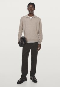 Man i beige långärmad polo, svarta byxor och svarta skor, håller en svart läderjacka mot en enkel vit bakgrund.