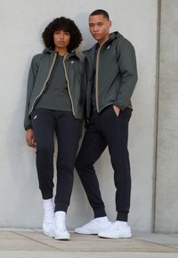 Giacca a vento verde con zip gialle e bianche, abbinata a pantaloni scuri e sneakers bianche. Il modello posa contro un muro di cemento.