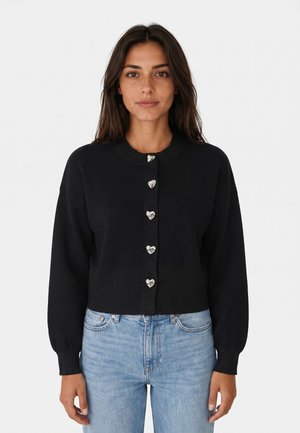 Donna che indossa un cardigan nero con bottoni a forma di cuore color argento e jeans azzurri, in piedi davanti a uno sfondo chiaro semplice.