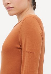 Long-sleeve top in earthy orange wool blend. Features a round neckline, flat seams, and subtle logo on the left sleeve. Smooth, fitted design.