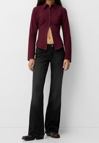 Camicia button-up fitted color bordeaux con maniche lunghe e design cropped, abbinata a jeans neri a gamba larga e stivali scuri.