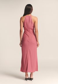 Rankovių suknelė iki pat grindų švelniai rožinės spalvos, su aukštu atvira nugara ir subtiliu šonu plyšiu. Lygus audinys su prigludusiu siluetu.