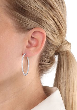Nahaufnahme eines Frauenohres mit einem großen silbernen Creolenohrring, zurückgebundenem blonden Haar und einem weißen Hemd mit Kragen.