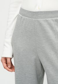 Κοντινό πλάνο ατόμου που φοράει ανοιχτόγριμο υφασμάτινο παντελόνι και λευκό μακρυμάνικο τοπ, με το χέρι χαλαρό δίπλα στο σώμα.