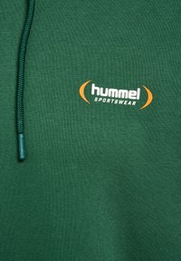 Dunkelgrüner Stoff mit geflochtenem Kordelzug und einem weißen und orangen "hummel SPORTSWEAR"-Logo, das darauf gedruckt ist.