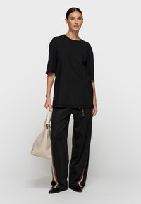 Mujer de pie con una camiseta negra holgada, pantalones negros con franjas laterales marrones y blancas, zapatos negros y sosteniendo un bolso beige.