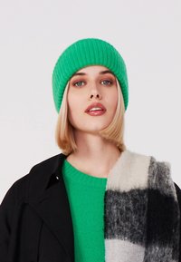 Groene geribbelde gebreide beanie, gecombineerd met een groene trui en een zwarte jas. Een grijs-wit geblokt sjaaltje voegt textuur en contrast toe.