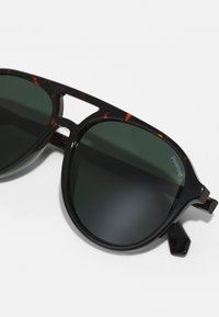Ochelari de soare cu ramă din fildes cu lentile verzi, având un design unic al punții și rame groase. Logo-ul Polaroid este vizibil pe lentilă.