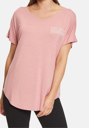 Kobieta w luźnym różowym T-shircie z krótkim rękawem i białym nadrukiem z motywem motyli na górnej lewej części klatki piersiowej.