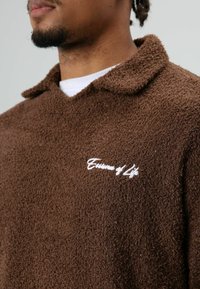 Polaire marron à col avec un texte brodé en blanc "Essence of Life." Texture douce, coupe ample, et détails de couture visibles.