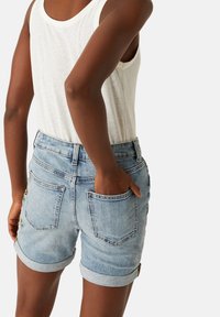 Shorts di denim blu chiaro con orli arrotolati, dotati di tasche posteriori e accenti ricamati colorati su un lato. Abbinati a un top canotta bianco.