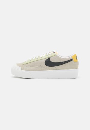 Baskets basses décontractées avec tige en daim beige, semelle blanche épaisse, logo Nike noir, lacets blancs et languette de talon jaune avec logo.