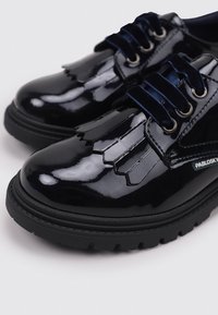 Zapatos de charol negros con puntera redondeada, que presentan recortes decorativos y cordones azul marino. Suela de goma negra resistente.